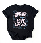 Flat lay of a Baking Is My Love Language shirt with baking tools and dessert graphics on black t-shirt on a white background