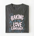 Flat lay of a Baking Is My Love Language shirt with baking tools and dessert graphics on charcoal t-shirt on a white background