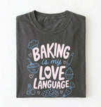 Flat lay of a Baking Is My Love Language shirt with baking tools and dessert graphics on charcoal t-shirt on a white background