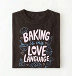Flat lay of a Baking Is My Love Language shirt with baking tools and dessert graphics on brown t-shirt on a white background