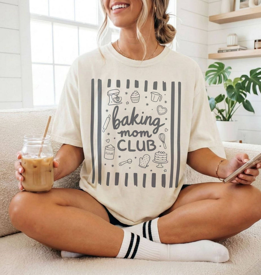 A woman sitting cross-legged on a couch wearing a cream-colored graphic t-shirt that reads 'baking mom CLUB' with baking-themed illustrations. She is holding an iced coffee in a glass jar and looking at her phone.