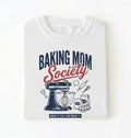 Flat lay of a white baking mom society shirt with stand mixer and baking ingredients baking mom gift on a white background 