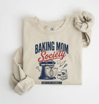 Flat lay of a sand Baking Mom Society graphic sweatshirt featuring a vintage mixer and baking tools for home bakers on a white background 