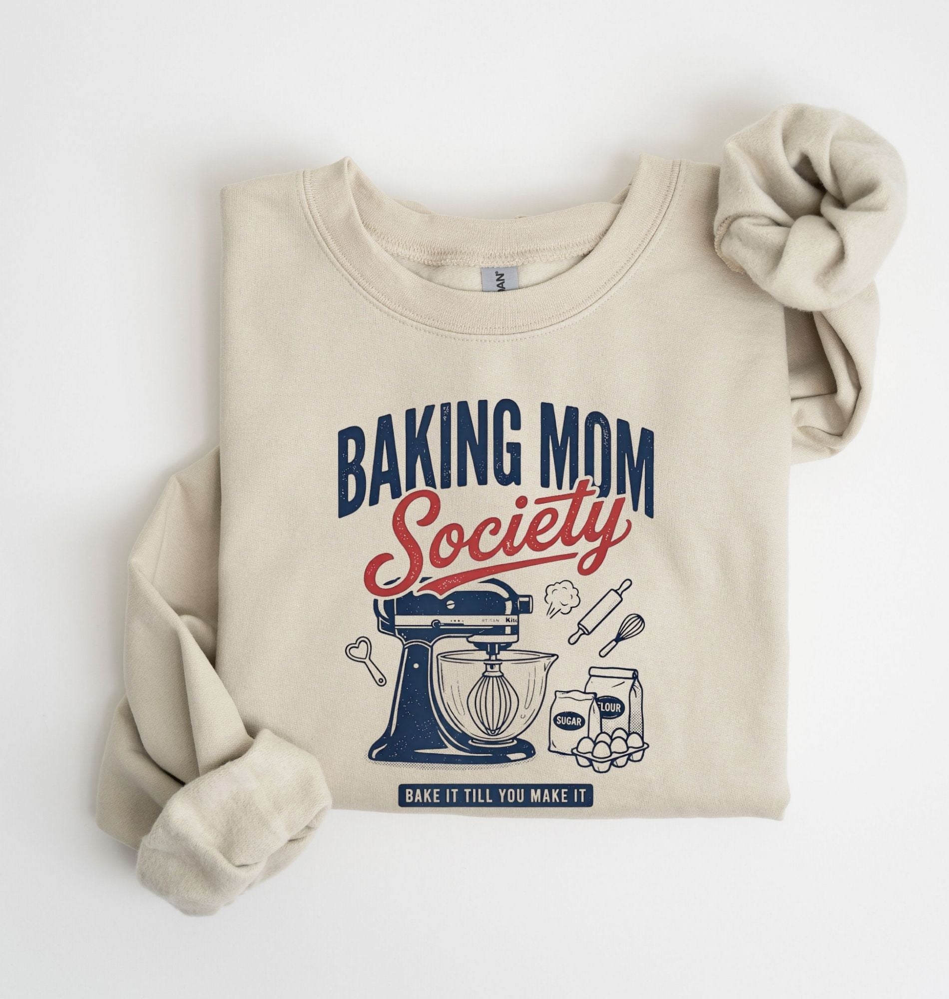 Flat lay of a sand Baking Mom Society graphic sweatshirt featuring a vintage mixer and baking tools for home bakers on a white background 