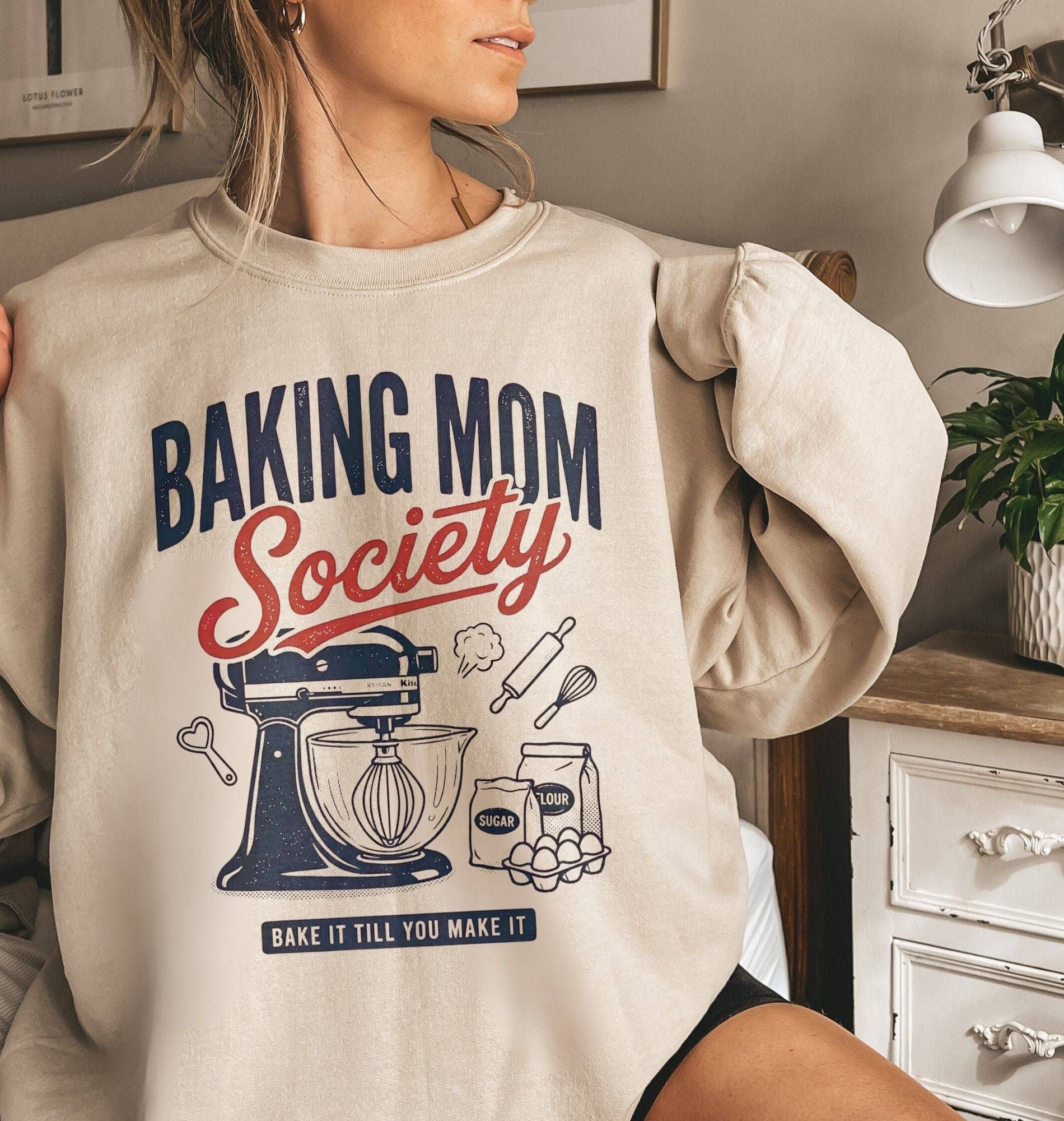A woman wearing a beige crewneck sweatshirt with a 'Baking Mom Society' graphic print in navy blue and red, featuring illustrations of a stand mixer, baking soda, and eggs. The photo is taken in a cozy, warm-toned room with a white dresser and lamp in the background.