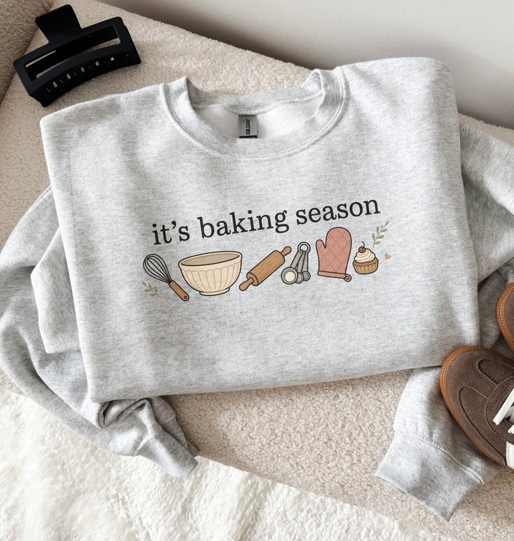 A folded light heather gray sweatshirt with a 'it's baking season' graphic featuring baking tools and treats, styled on a cream textured blanket with a black hair clip, brown shoes, and a small dish in the background.