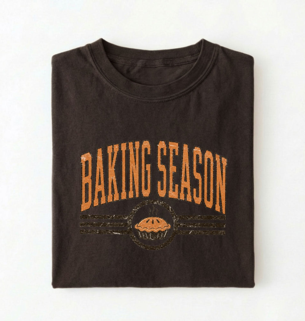 A dark brown folded t-shirt with a distressed orange graphic that reads 'BAKING SEASON' above a pie illustration, displayed on a clean white background.