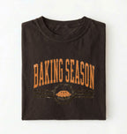A dark brown folded t-shirt with a distressed orange graphic that reads 'BAKING SEASON' above a pie illustration, displayed on a clean white background.
