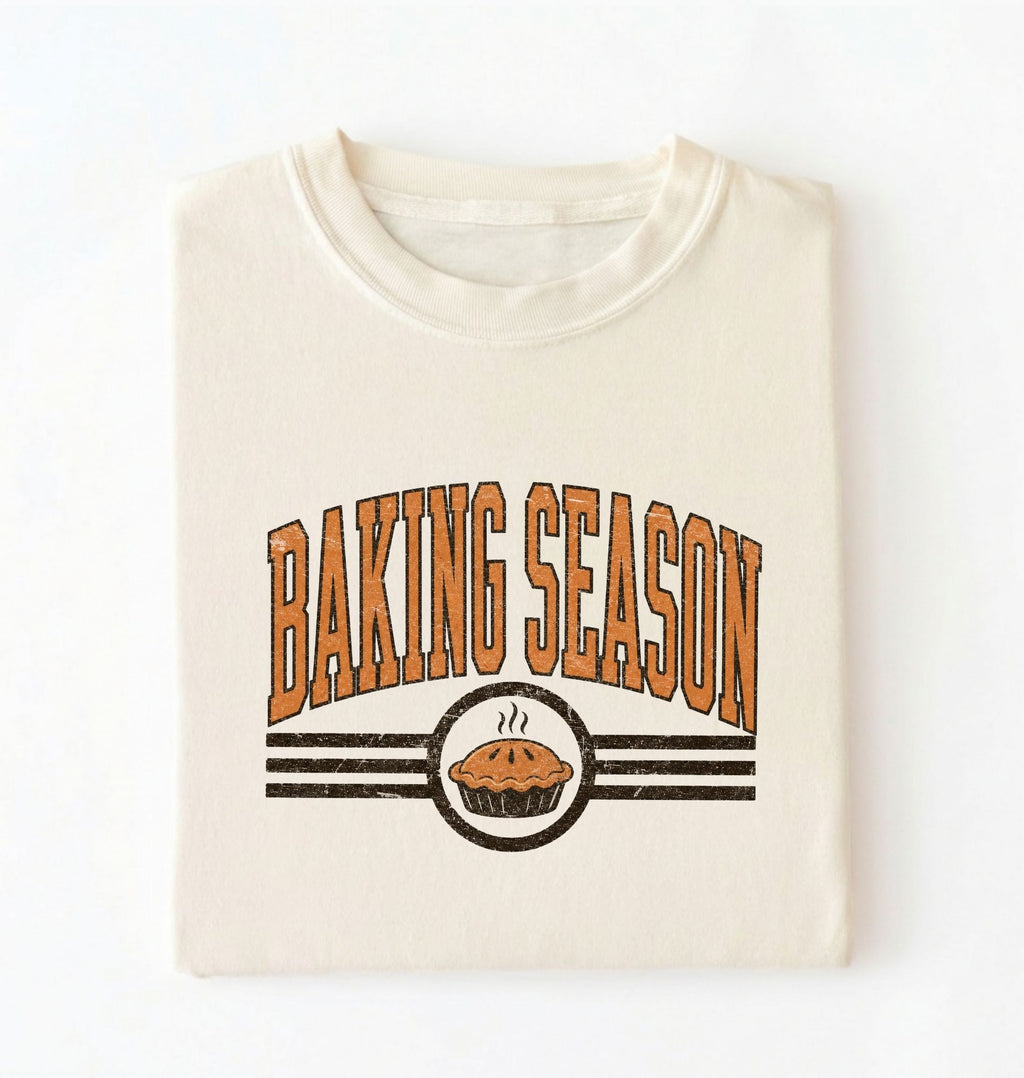 A folded cream-colored t-shirt with a retro graphic that reads 'BAKING SEASON' in orange-brown letters above a pie illustration, set against a plain white background.
