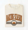 A folded cream-colored t-shirt with a retro graphic that reads 'BAKING SEASON' in orange-brown letters above a pie illustration, set against a plain white background.
