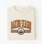 A folded cream-colored t-shirt with a retro graphic that reads 'BAKING SEASON' in orange-brown letters above a pie illustration, set against a plain white background.