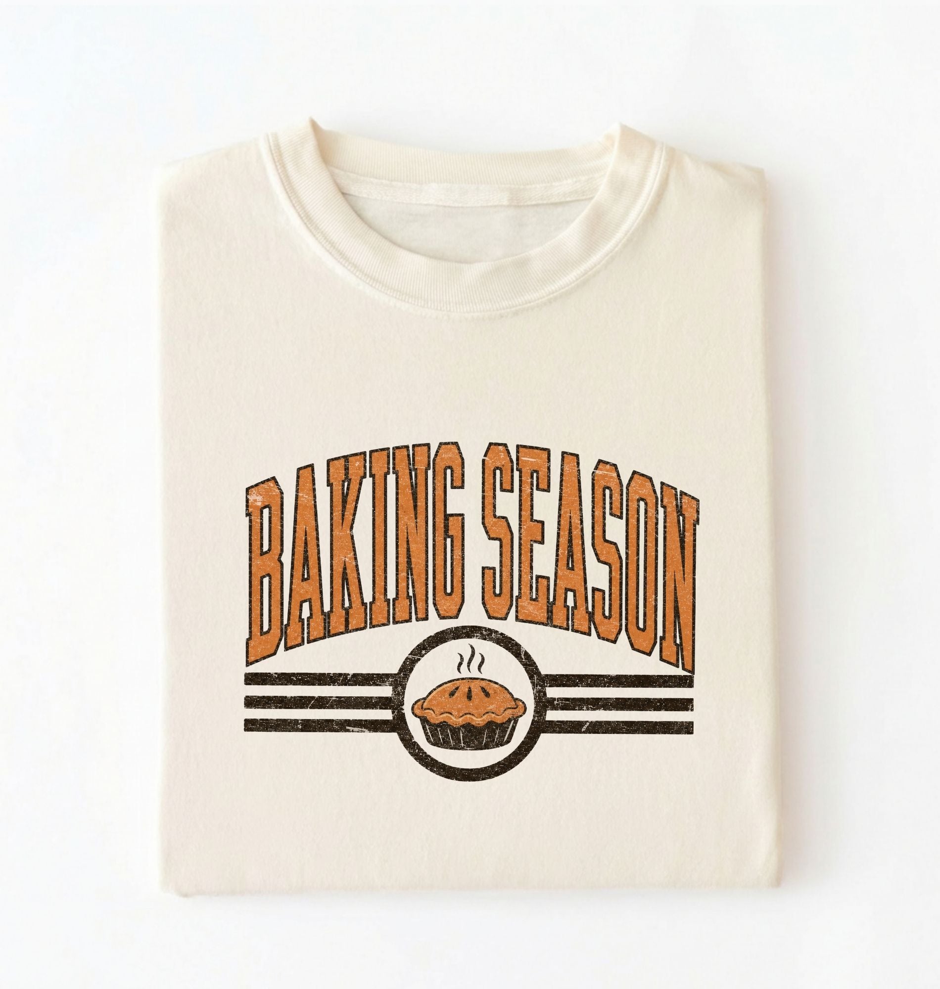 A folded cream-colored t-shirt with a retro graphic that reads 'BAKING SEASON' in orange-brown letters above a pie illustration, set against a plain white background.