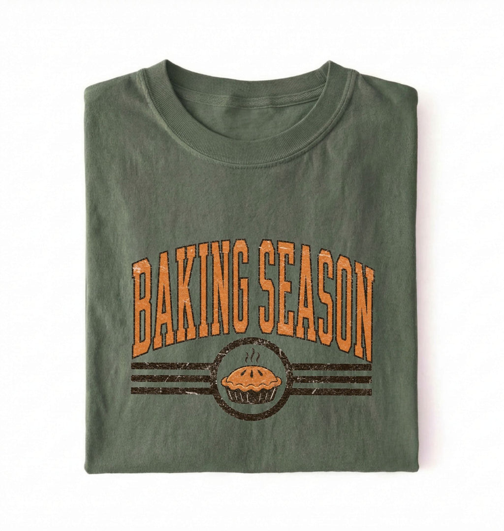 A folded olive green t-shirt with a distressed orange and brown graphic that reads 'BAKING SEASON' above an illustration of a pie, set against a plain white background.