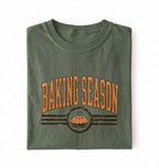 A folded olive green t-shirt with a distressed orange and brown graphic that reads 'BAKING SEASON' above an illustration of a pie, set against a plain white background.