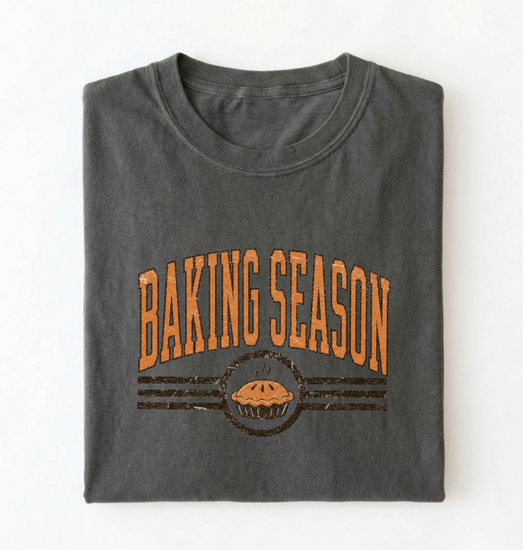 A folded dark gray t-shirt with a distressed orange graphic that reads 'BAKING SEASON' above an illustration of a pie, set against a plain white background.
