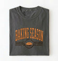 A folded dark gray t-shirt with a distressed orange graphic that reads 'BAKING SEASON' above an illustration of a pie, set against a plain white background.