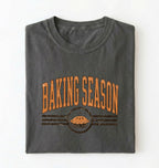A folded dark gray t-shirt with a distressed orange graphic that reads 'BAKING SEASON' above an illustration of a pie, set against a plain white background.