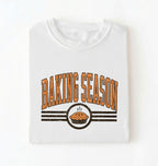 A folded white t-shirt with a retro-style graphic that reads 'BAKING SEASON' in orange arched letters above a pie illustration and horizontal stripe on a white background 
