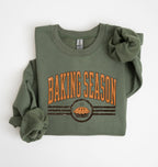 An olive green crewneck sweatshirt with rolled sleeves, featuring a distressed orange and brown graphic that reads 'BAKING SEASON' above a cupcake illustration, laid flat on a white background.
