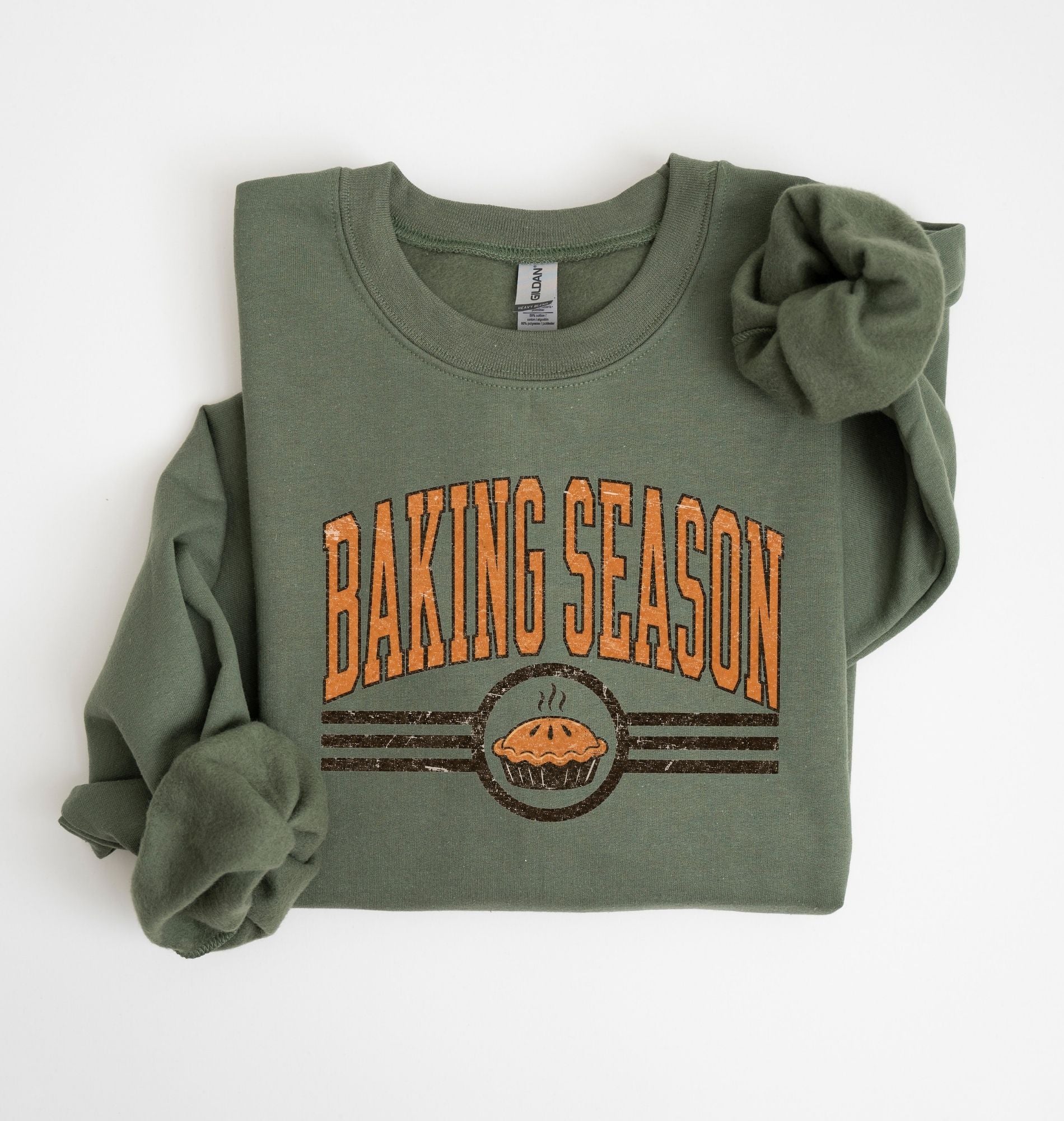 An olive green crewneck sweatshirt with rolled sleeves, featuring a distressed orange and brown graphic that reads 'BAKING SEASON' above a cupcake illustration, laid flat on a white background.