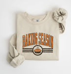 A folded beige crewneck sweatshirt with rolled sleeves, featuring a retro-style graphic that reads 'BAKING SEASON' in orange arched letters above a pie illustration, on a clean white background.