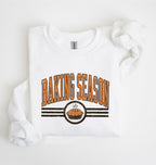 A white long-sleeve sweatshirt laid flat on a white background, featuring a graphic print that reads 'BAKING SEASON' in orange arched letters above a pie illustration and dark horizontal stripes.