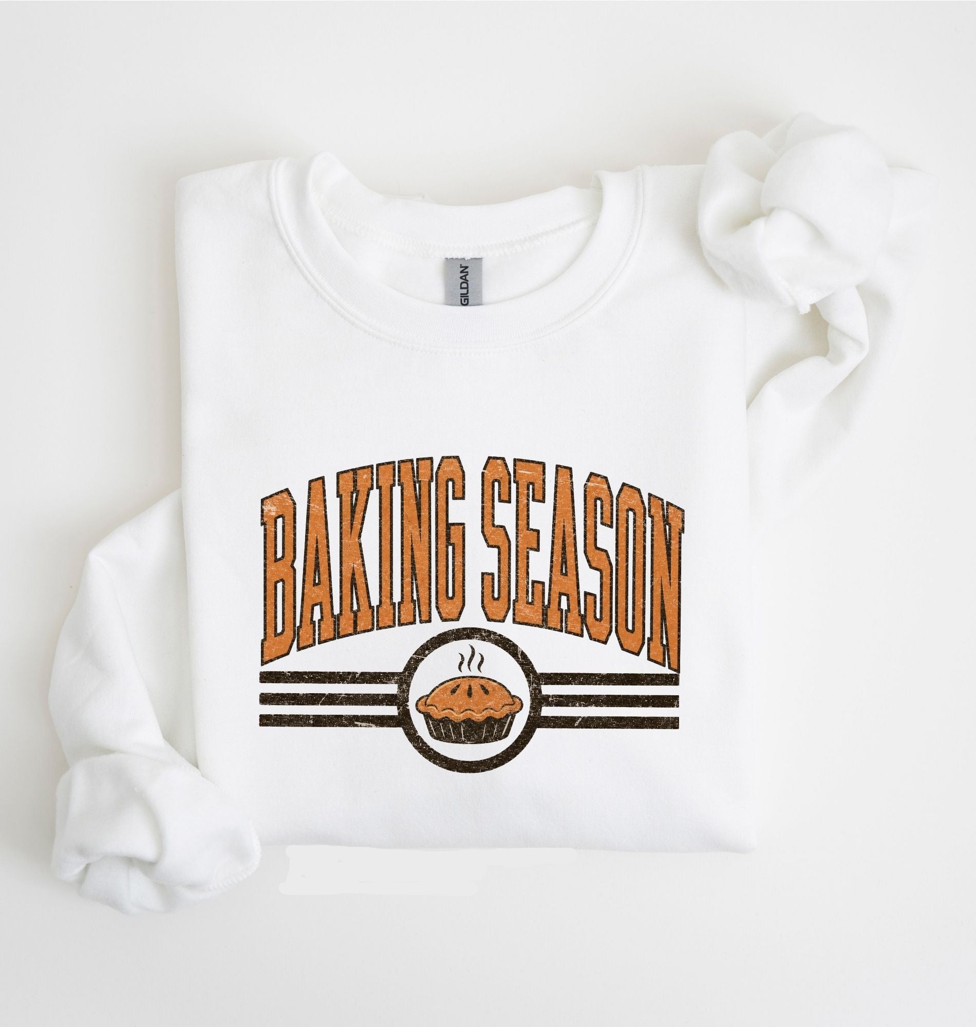 A white long-sleeve sweatshirt laid flat on a white background, featuring a graphic print that reads 'BAKING SEASON' in orange arched letters above a pie illustration and dark horizontal stripes.