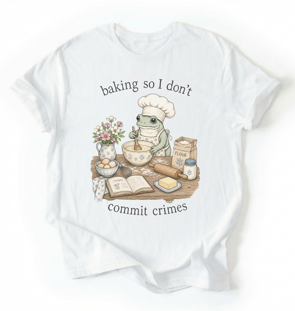A white short-sleeve t-shirt with a graphic of a green frog wearing a chef's hat and apron, mixing batter at a baking table with ingredients like flour, eggs, and butter. The text reads 'baking so I don't commit crimes'.