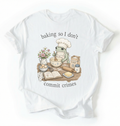 A white short-sleeve t-shirt with a graphic of a green frog wearing a chef's hat and apron, mixing batter at a baking table with ingredients like flour, eggs, and butter. The text reads 'baking so I don't commit crimes'.