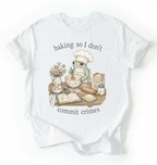 A white short-sleeve t-shirt with a graphic of a green frog wearing a chef's hat and apron, mixing batter at a baking table with ingredients like flour, eggs, and butter. The text reads 'baking so I don't commit crimes'.