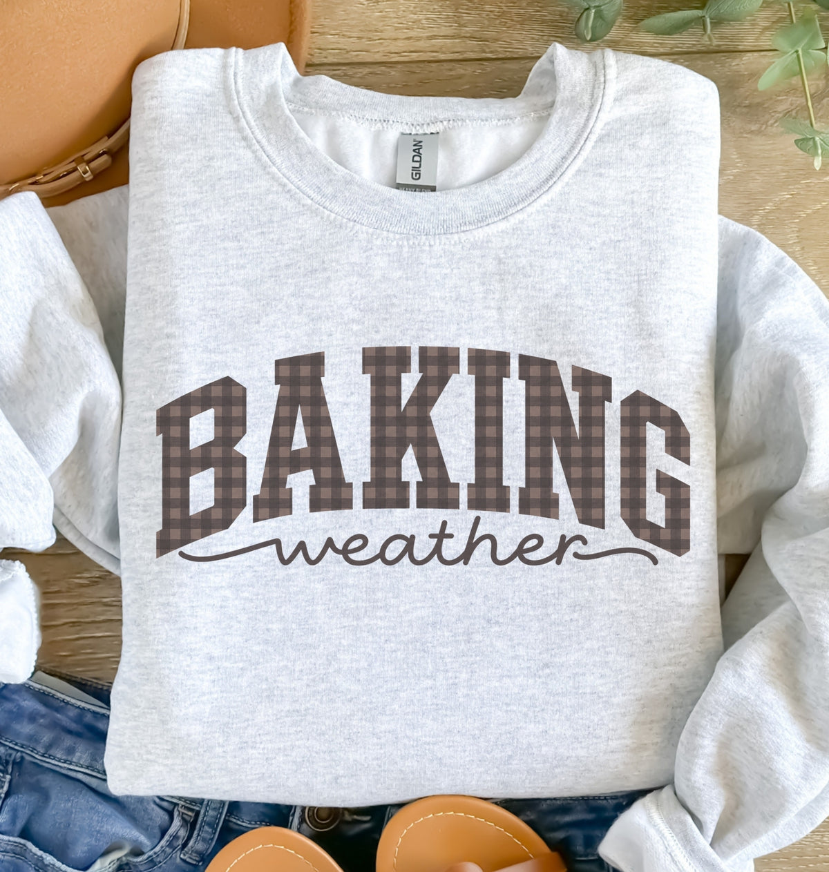 An ash crewneck sweatshirt with 'BAKING weather' printed on the front. The word 'BAKING' is in a brown and black plaid pattern, while 'weather' is in black script.