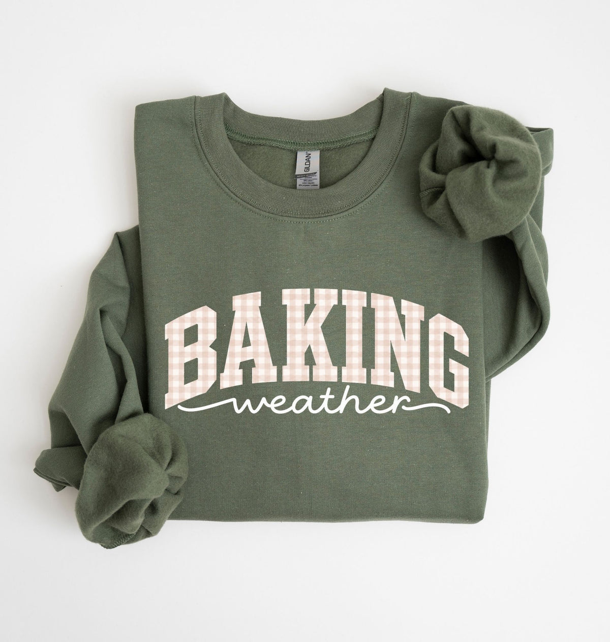 An olive green long-sleeve sweatshirt with 'BAKING weather' printed on the front in white and pink gingham, styled flat on a white background with a matching scrunchie.