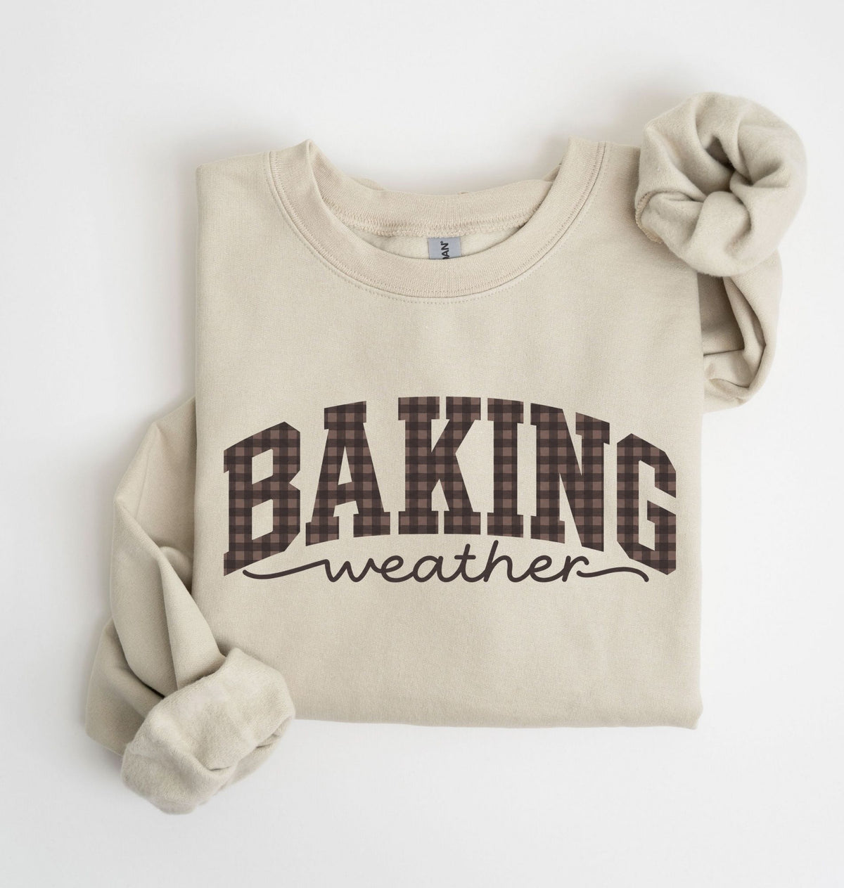 A beige crewneck sweatshirt with 'BAKING weather' printed on the front. The word 'BAKING' is in a brown and black plaid pattern, while 'weather' is in black script.