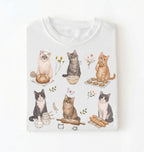 White t-shirt with cat illustrations and baking items on a white background