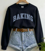 A black long-sleeve crewneck sweatshirt with 'BAKING' printed in white varsity-style letters across the chest, styled on a wooden hanger with light-wash denim shorts, a brown belt, and a small green plant in the corner.