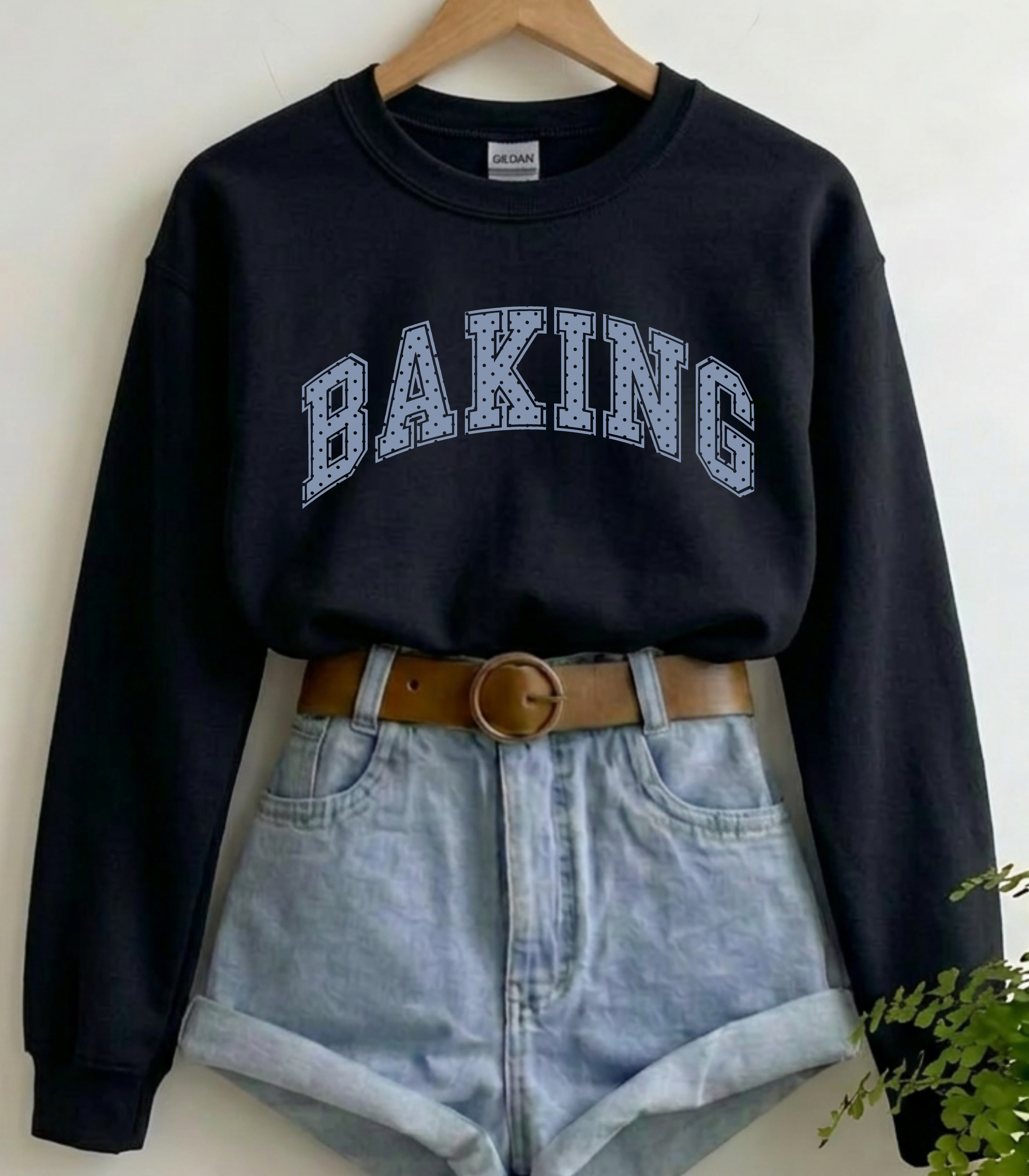 A black long-sleeve crewneck sweatshirt with 'BAKING' printed in white varsity-style letters across the chest, styled on a wooden hanger with light-wash denim shorts, a brown belt, and a small green plant in the corner.