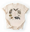 Flat lay of a natural Cottagecore Be Kind To All Kinds shirt with woodland animals illustration on a white background