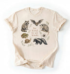 Flat lay of a natural Cottagecore Be Kind To All Kinds shirt with woodland animals illustration on a white background
