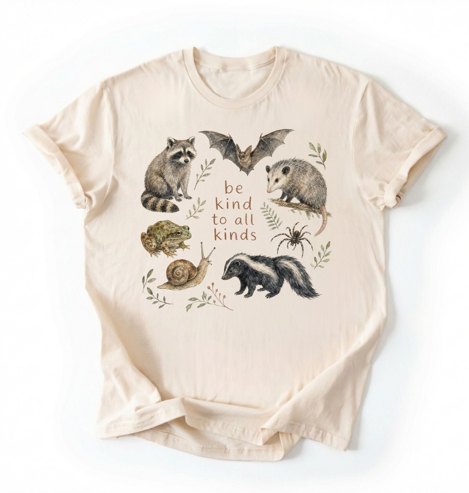 Flat lay of a natural Cottagecore Be Kind To All Kinds shirt with woodland animals illustration on a white background