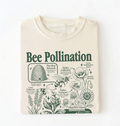 A natural crewneck t-shirt with a detailed green graphic print about bee pollination, featuring illustrations of a beehive, flowers, bees, and pollen transport, laid flat against a white background.