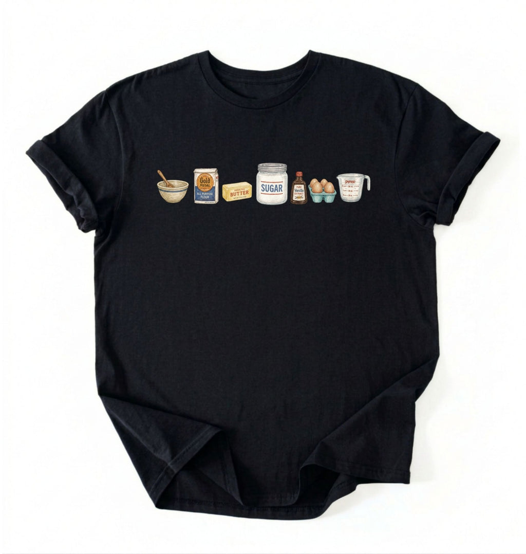 A black short-sleeve t-shirt with a graphic print of baking ingredients (a bowl, flour bag, butter, sugar jar, vanilla extract bottle, eggs, and milk carton) lined up across the chest, displayed flat against a white background.