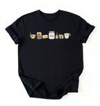 A black short-sleeve t-shirt with a graphic print of baking ingredients (a bowl, flour bag, butter, sugar jar, vanilla extract bottle, eggs, and milk carton) lined up across the chest, displayed flat against a white background.