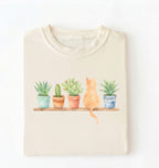 Flat lay of a ivory t-shirt featuring a watercolor illustration of potted plants on a shelf with a cat sitting among them on a white background 