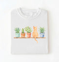 Flat lay of a white t-shirt featuring a watercolor illustration of potted plants on a shelf with a cat sitting among them on a white background 