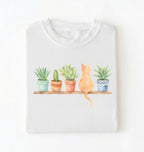 Flat lay of a white t-shirt featuring a watercolor illustration of potted plants on a shelf with a cat sitting among them on a white background 