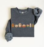 A dark heather gray crewneck sweatshirt with long sleeves, laid flat on a light cream background. The front of the sweatshirt features a horizontal row of colorful Christmas-themed food illustrations, including a gingerbread man, a bowl, a jar of peanut butter, a pie, eggs, hot chocolate, and a Christmas tree.