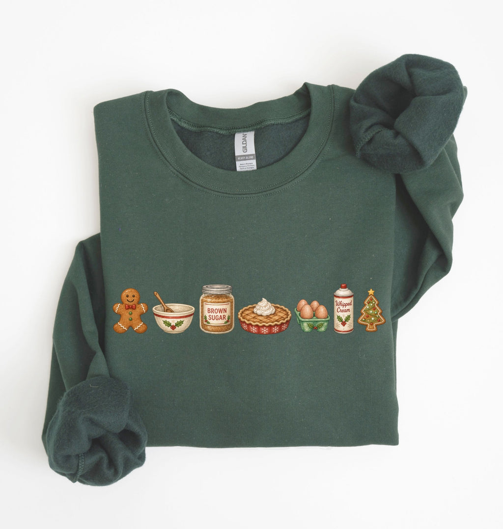 A dark green crewneck sweatshirt with a holiday-themed food line graphic on the chest, styled flat on a white background with a matching green knit scrunchie.