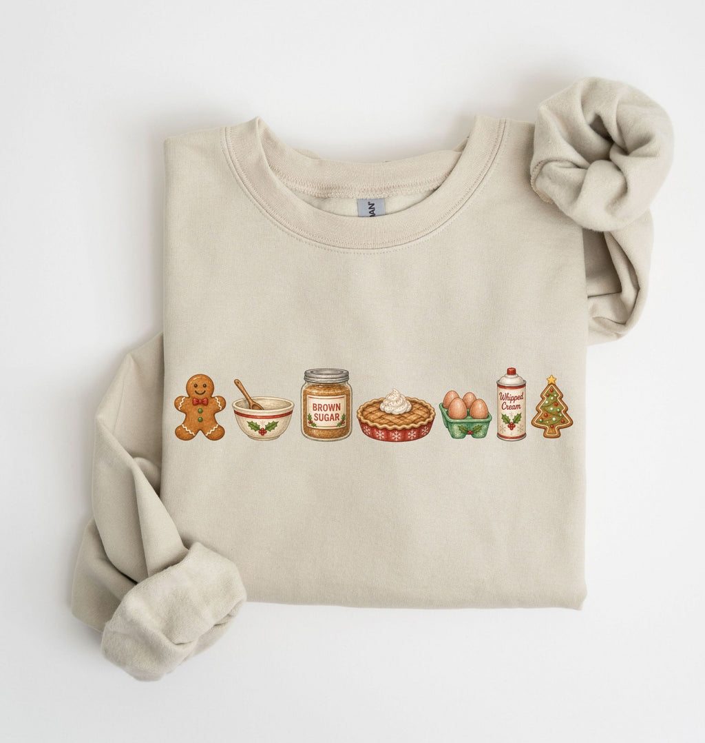 A beige crewneck sweatshirt with a holiday-themed graphic of gingerbread cookies, a bowl, a jar of brown sugar, a pie, eggs, milk, and a Christmas tree. The sweatshirt is styled flat on a white background with one sleeve rolled up and a matching fabric scrunchie.