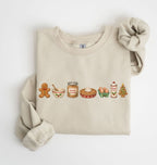 A beige crewneck sweatshirt with a holiday-themed graphic of gingerbread cookies, a bowl, a jar of brown sugar, a pie, eggs, milk, and a Christmas tree. The sweatshirt is styled flat on a white background with one sleeve rolled up and a matching fabric scrunchie.