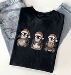 A folded black t-shirt with a graphic of three penguins wearing Santa hats and sitting in winter clothing, styled on top of light-wash blue jeans against a white background.